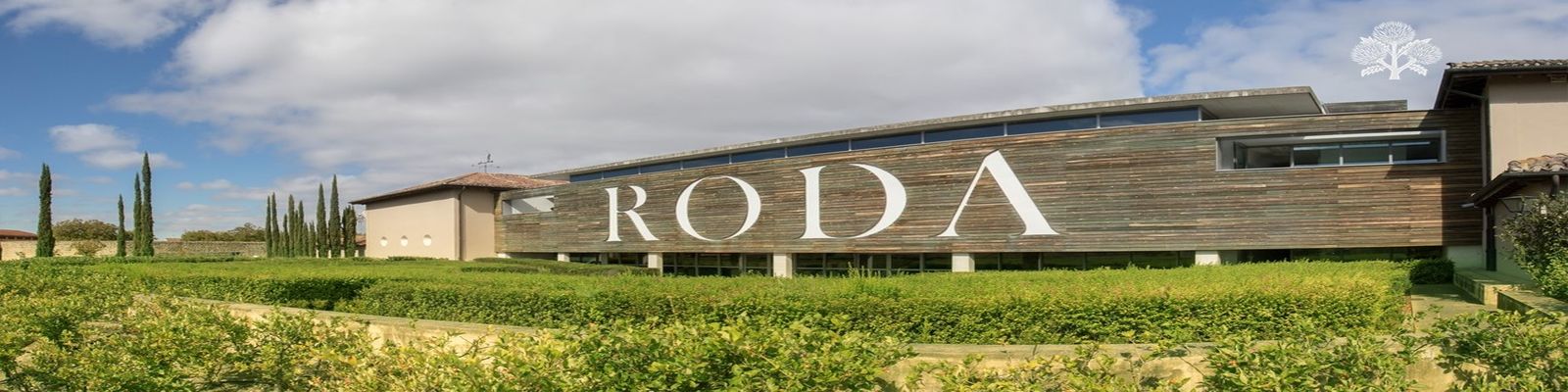 Bodegas Roda Ein modernes Gebäude mit einer Holzfassade, auf der der Schriftzug -RODA- in großen Buchstaben zu sehen ist. Umgeben von grünen Weinreben und Zypressen unter einem bewölkten Himmel.