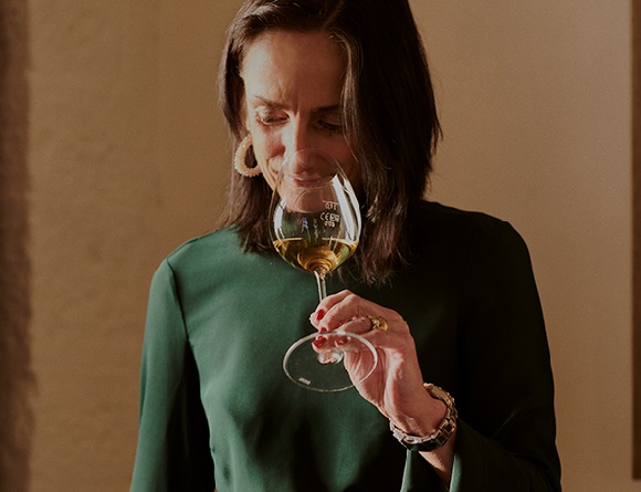 Eine Frau mit schulterlangen Haaren trägt ein grünes Oberteil und schaut nachdenklich auf ein Weinglas, während sie den Wein untersucht. Der Hintergrund ist unscharf und hell.