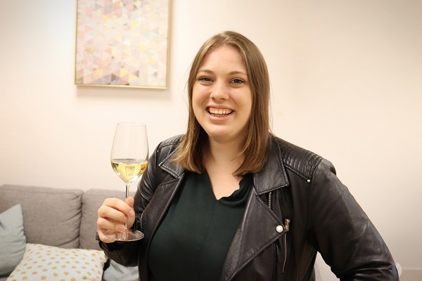 Eine junge Frau lächelt und hält ein Glas Wein in der Hand. Sie trägt eine schwarze Lederjacke und steht in einem hellen Raum mit einem Sofa und einem Kunstwerk an der Wand.