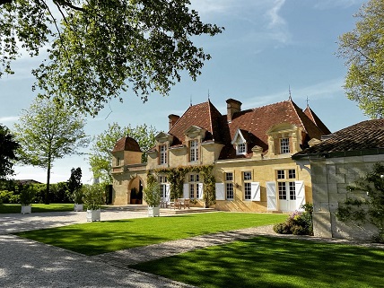 Ein beeindruckendes, elegantes Herrenhaus mit roten Dächern und hellen Wänden steht im Mittelpunkt, umgeben von gepflegtem Gras und mehreren Bäumen an einem sonnigen Tag.