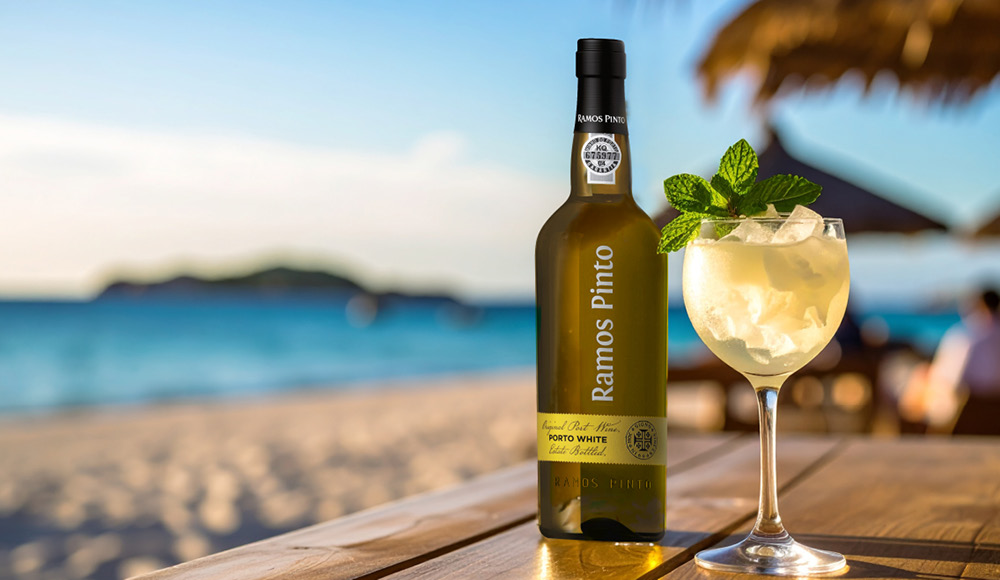 Eine Flasche Ramos Pinto Porto White steht auf einem Tisch am Strand, daneben ein Glas mit Eiswürfeln und Minze. Im Hintergrund ist das Meer und ein sonniger Himmel sichtbar.