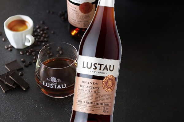 SDM_November_600 x 400 Eine Flasche Brandy de Jerez steht im Vordergrund, daneben ein Glas mit dunklem Likör. Im Hintergrund sind eine Tasse Kaffee und Kaffeebohnen sowie eine weitere Flasche zu sehen. Dunkle Schokoladentafeln liegen ebenfalls auf der Oberfläche.