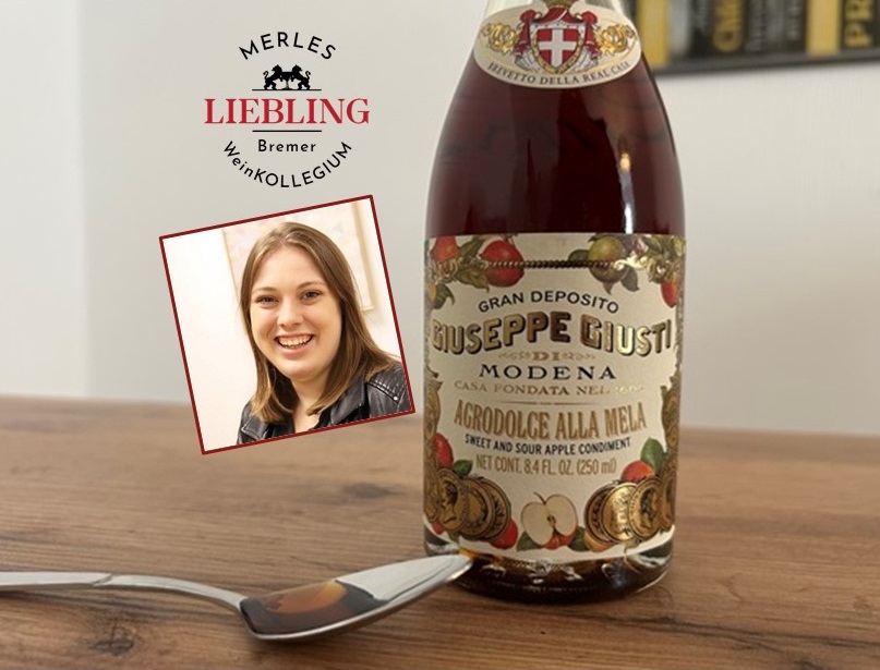 Ein Glasbehälter mit einer etikettierten Flasche -Giuseppe Giusti- steht auf einem Tisch. Ein Löffel ist daneben positioniert. Im Hintergrund ist der Schriftzug -MERLES LIEBLING Bremer WeinKOLLEGIUM- und ein eingefügtes Bild einer Person zu sehen.