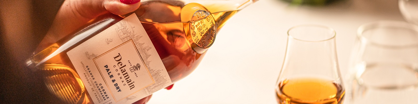 Cognac Delamain Eine Person hält eine Flasche Cognac mit einem eleganten Etikett und gießt ihn in ein Glas. Im Hintergrund sind unscharfe Gläser und eine helle Umgebung sichtbar.