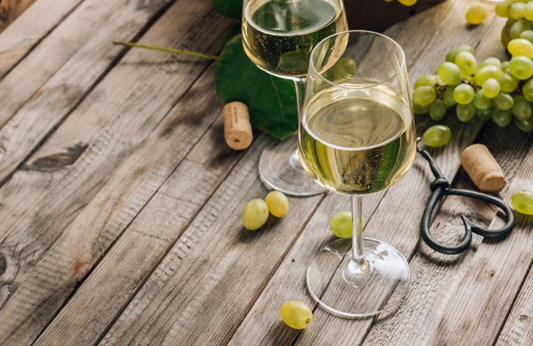 Zwei Gläser mit Weißwein stehen auf einem Holztablett. Um sie herum liegen Trauben, Korken und ein Korkenzieher. Die Umgebung wirkt gemütlich und rustikal.