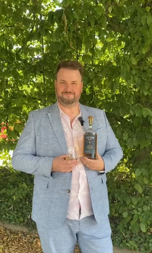 Ein Mann in einem hellblauen Anzug steht vor einem grünen Hintergrund und hält eine Flasche Spirituose sowie ein Glas. Er lächelt, während er die Objekte präsentiert.
