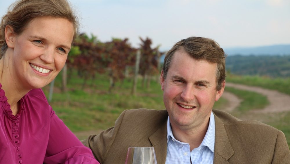 Auf einen Wein mit…Prinz Felix zu Salm-Salm, Weingut Prinz Salm Ein Paar lächelt in einem Weinberg, während sie an einem Tisch sitzen. Die Frau trägt ein lila Oberteil, der Mann einen braunen Anzug. Im Hintergrund sind Weinreben und ein Weg sichtbar.