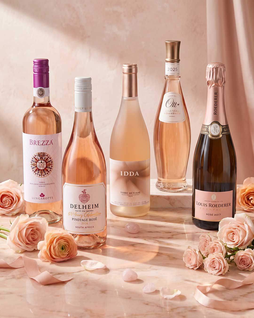 Fünf Flaschen Roséwein stehen auf einem hellen Marmortisch, umgeben von rosa Rosen und Blütenblättern. Der Hintergrund ist sanft und neutral gehalten, was die Farben der Weine betont.