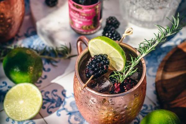 Ein kupferner Becher enthält ein Getränk mit Brombeeren, Limettenscheiben und einem Rosmarinzweig. Im Hintergrund sind grüne Limetten und eine Flasche mit Sirup zu sehen.