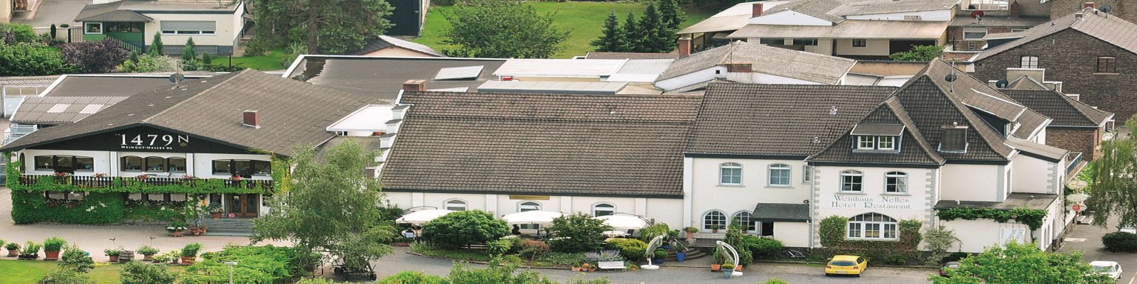 Nelles Ein Hotelgebäude mit einem einladenden Außenbereich, Tischen und Stühlen unter Sonnenschirmen. Umgeben von anderen Gebäuden und Grünflächen, steht ein gelbes Auto auf dem Parkplatz.