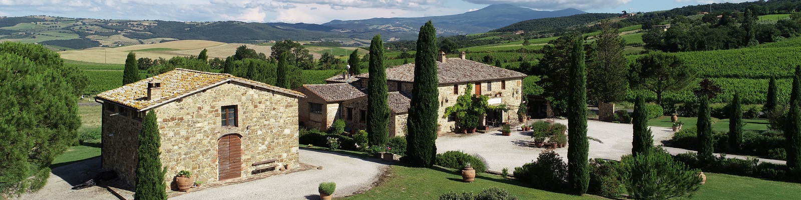 Casanova di Neri Ein steinernes Landhaus mit einem angrenzenden Nebengebäude steht in einer malerischen ländlichen Umgebung. Die Umgebung ist von grünen Hügeln und Weinbergen geprägt, unter einem blauen Himmel mit Wolken.
