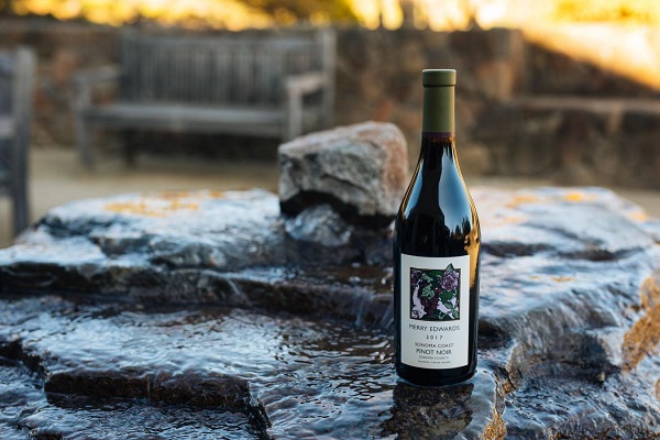 Eine Weinflasche steht auf einem steinernen Wasserspiel, während Wasser darüber fließt. Im Hintergrund sind eine Bank und eine rustikale Umgebung sichtbar.