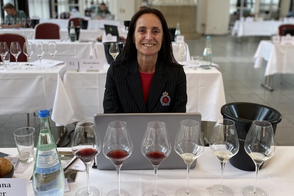 Eine Person sitzt vor einem Laptop, umgeben von mehreren Weingläsern mit Rot- und Weißwein. Im Hintergrund sind Tische mit weiteren Gläsern und Wasserflaschen zu sehen, in einem hellen Veranstaltungsraum.