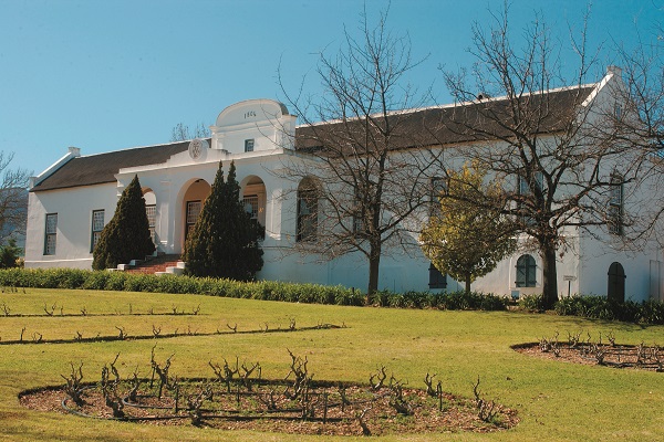 Ein historisches, weißes Gebäude mit einem großen Eingangsbereich und rundem Dach steht in einer grünen Landschaft. Vor dem Gebäude sind kahle Bäume und ein kleiner Garten mit Pflanzen angelegt.