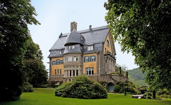 Eine große Villa steht inmitten von Grünanlagen, umgeben von hohen Bäumen und Sträuchern. Die Architektur wirkt historisch, mit Türmen und großen Fenstern, die auf eine entspannte Landschaft blicken.