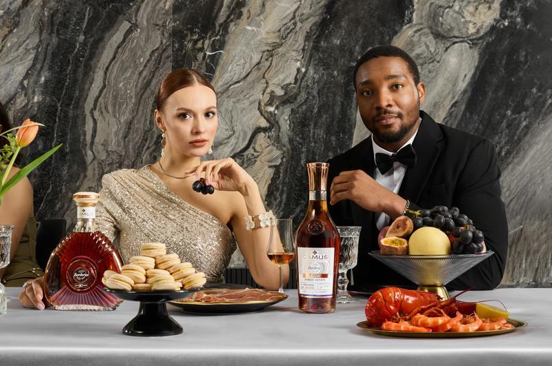 Ein elegantes Paar sitzt an einem Tisch, umgeben von Luxusessen wie Macarons, Meeresfrüchten und verschiedenen Früchten, während sie Getränke genießen und in die Kamera blicken.