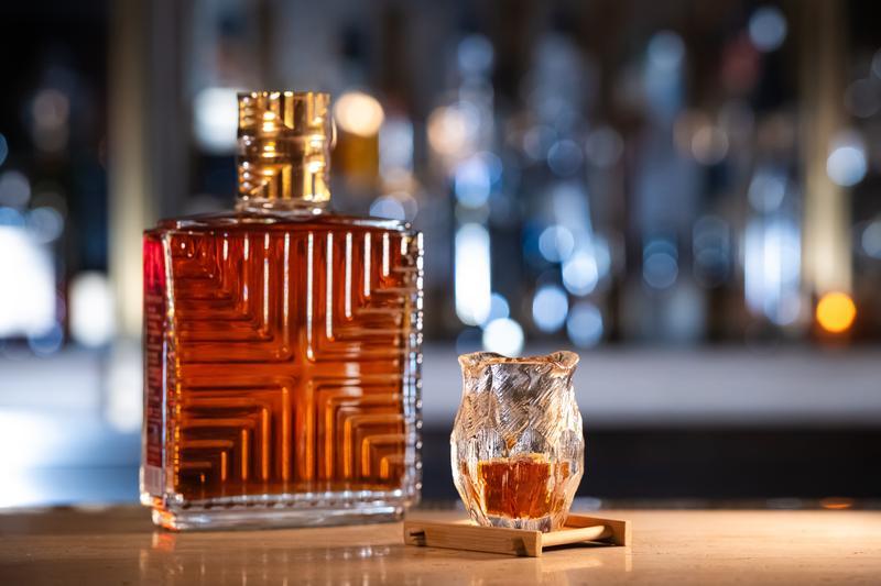 Ein eleganter, rechteckiger Flasche für alkoholisches Getränk steht neben einem kunstvoll gestalteten Glas auf einem kleinen Holzständer, umgeben von unscharfen Lichtern im Hintergrund.