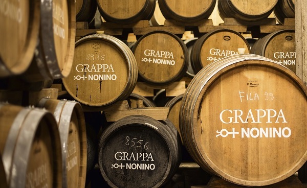 Holzfässer stehen gestapelt in einer Lagerhalle. Einige Fässer sind mit -GRAPPA NONINO- beschriftet, während andere Nummern und Jahrgänge zeigen. Das Licht ist warm und die Umgebung wirkt rustikal.