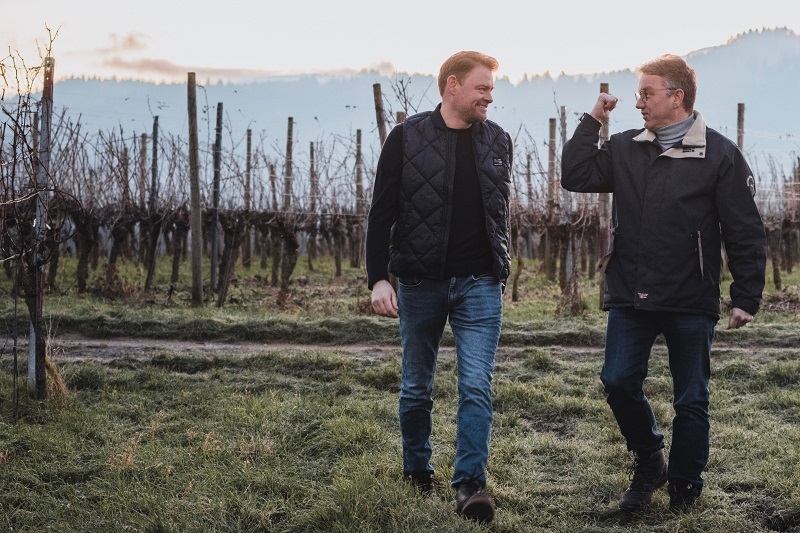 Zwei Männer unterhalten sich lächelnd und gehen durch einen Weinberg. Im Hintergrund sind Weinreben und eine sanfte Landschaft mit einem bewölkten Himmel zu sehen.
