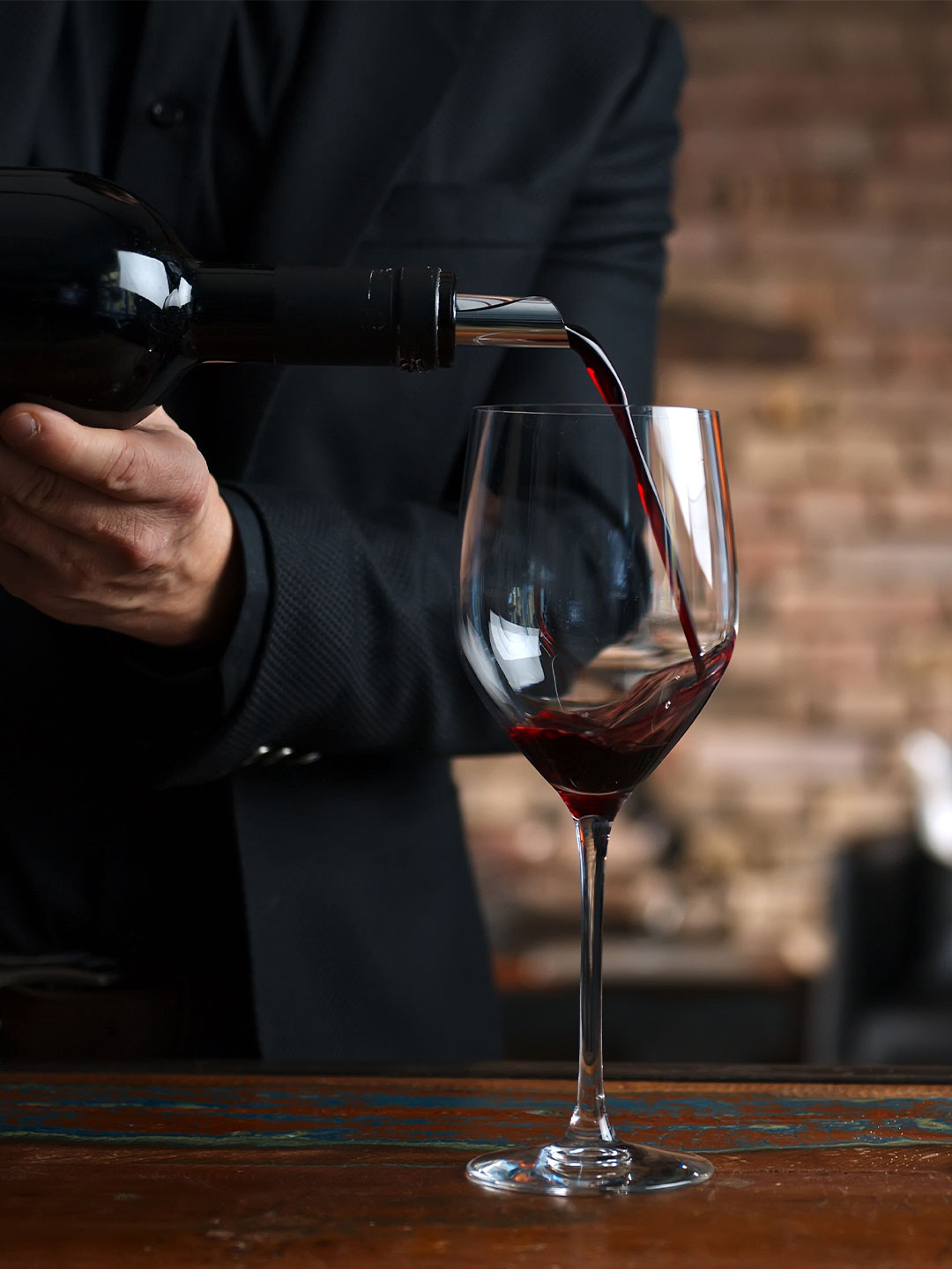 Ein Mann in einem schwarzen Anzug gießt Rotwein aus einer Flasche in ein schönes Weinglas, während er an einem holzfarbenen Tisch steht. Im Hintergrund sind unscharfe, rustikale Elemente sichtbar.