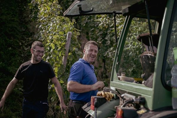 Zwei Männer lächeln und stehen neben einem Traktor, umgeben von Weinreben. Einer trägt ein blaues Hemd, der andere ein schwarzes. Es ist ein sonniger Tag im Freien.
