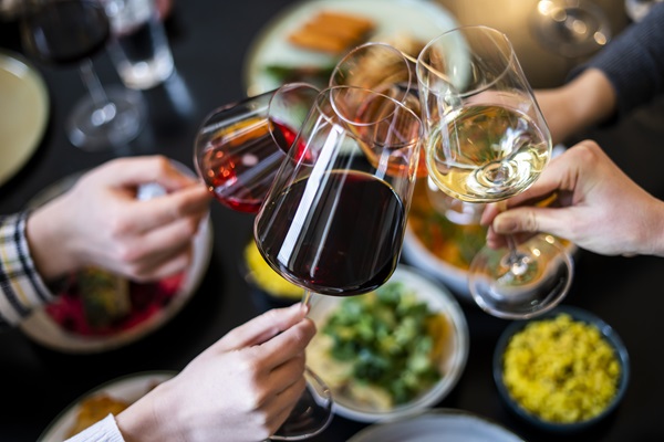 Verschiedene Hände stoßen mit Weingläsern an. Auf dem Tisch sind mehrere bunte, appetitlich angerichtete Gerichte zu sehen, darunter Salat, Reis und andere Speisen.