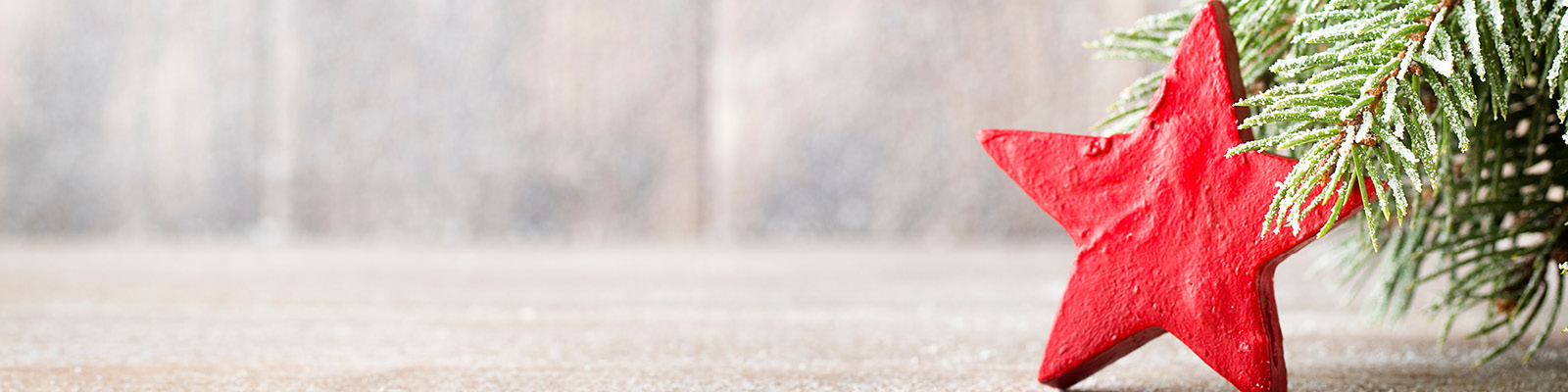 Ein roter Holzstern steht auf einer Holzoberfläche. Daneben ist ein grüner, mit Schnee bestäubter Zweig zu sehen. Im Hintergrund verschwommenes, neutrales Holz.