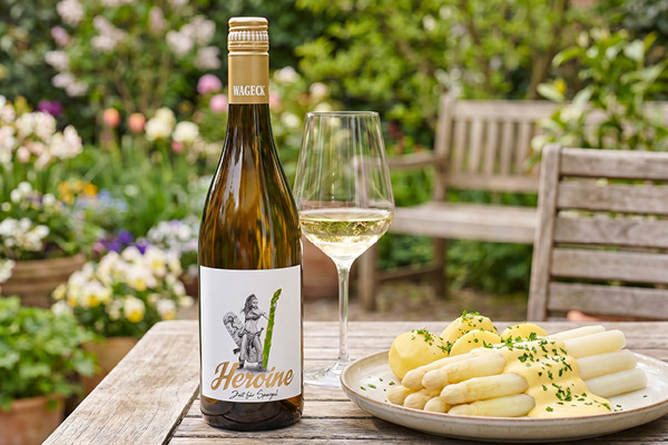 Eine Flasche Weißwein mit dem Etikett -Heroine- steht neben einem Glas Wein auf einem Garten Tisch. Auf einem Teller liegt Spargel mit Kartoffeln, umgeben von blühenden Pflanzen im Hintergrund.