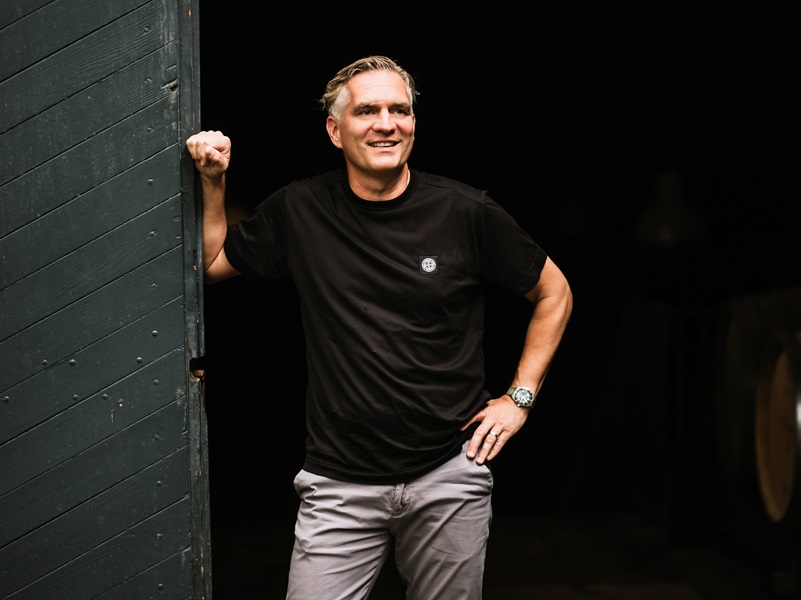 Ein Mann steht in einem Torbogen. Er trägt ein schwarzes T-Shirt und graue Hosen, lächelt und hat eine Hand an der Wand. Die Umgebung ist dunkel.