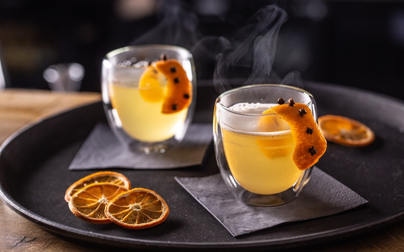 Ein dampfender Becher mit bräunlichem Getränk steht auf einem Beigen Tuch. Er ist mit einer orangenen Schale dekoriert, vor einem dunklen Hintergrund.