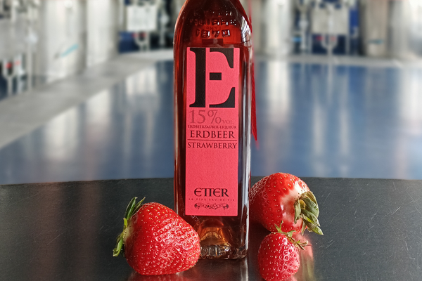 Eine Flasche Erdbeerlikör mit dem Aufdruck -E- und -15% vol- steht auf einer schwarzen Oberfläche. Neben der Flasche liegen frische Erdbeeren. Der Hintergrund ist unscharf und zeigt eine hellere industrielle Umgebung.