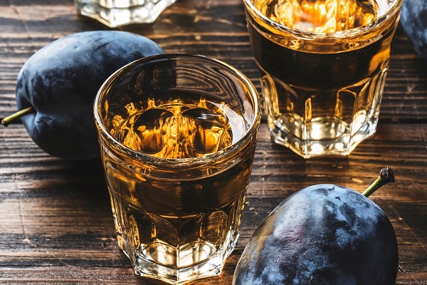 Zwei Gläser mit goldenem Getränk stehen auf einem dunklen Holzuntergrund, umgeben von frischen, blauen Pflaumen. Die Gläser haben Eiswürfel und reflektieren das Licht.