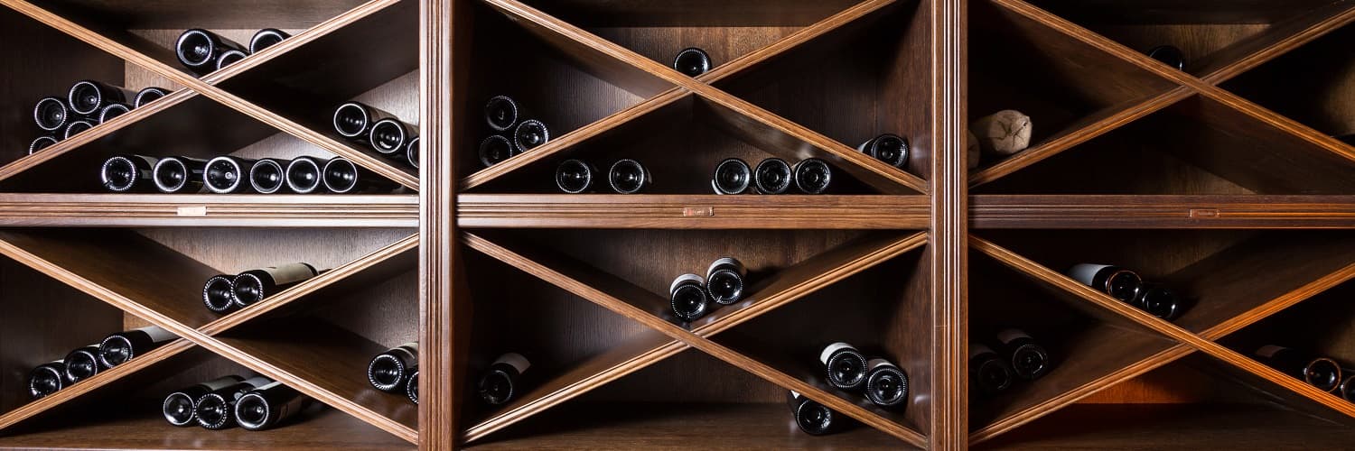 Wein lagern? Wir zeigen, wie es richtig geht! Ein Weinständer aus Holz mit diagonal angeordneten Fächern, in denen zahlreiche Weinflaschen liegen. Die Flaschen sind schwarz und teils gut sichtbar, während einige versteckt sind.
