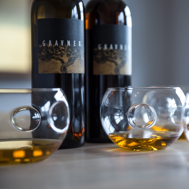 Zwei Flaschen Wein mit der Aufschrift -Gravner- stehen hinter mehreren bauchigen Gläsern. Die Gläser enthalten bernsteinfarbenen Wein und zeigen ein einzigartiges Design mit einem runden Ausschnitt.