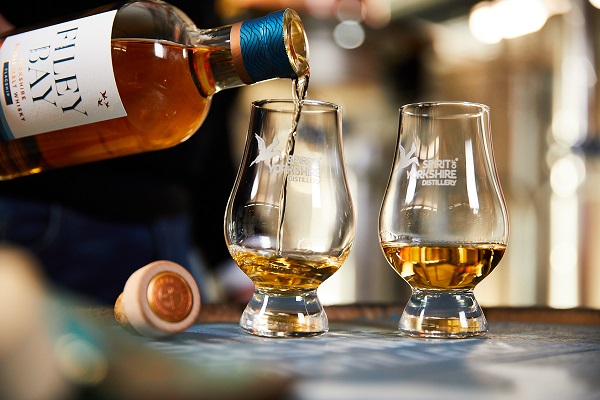 Eine Flasche Whisky wird ausgehoben und in zwei Gläser mit goldenem Inhalt eingegossen. Die Gläser sind mit einem Logo der -Spirit of Yorkshire Distillery- versehen. Der Hintergrund ist unscharf.