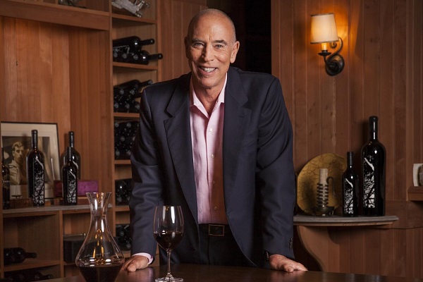 Ein Mann steht lächelnd an einem Tisch in einem Weinkeller. Er trägt einen dunkelblauen Anzug und hat ein Glas Rotwein in der Hand. Im Hintergrund sind Weinflaschen und dekorative Elemente sichtbar.