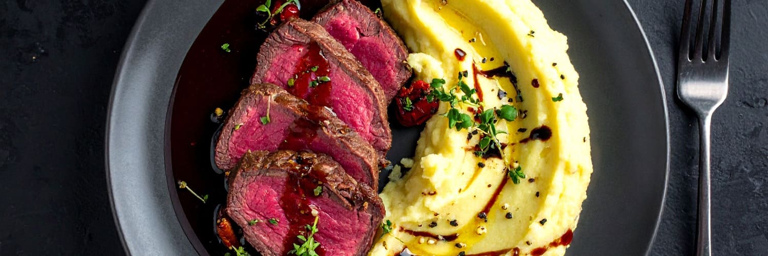 Ein Glas Rotwein steht im Vordergrund. Im Hintergrund liegt ein kunstvoll angerichteter Teller mit zwei Scheiben Fleisch in einer Sauce, umgeben von kleinen Beilagen und frischen Kräutern.