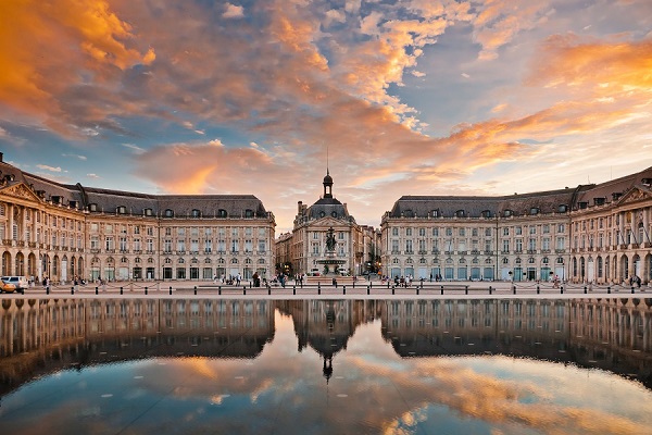 Ein eindrucksvoller Platz spiegelt sich im Wasser eines Brunnens wider. Historische Gebäude umgeben den Platz, während Menschen spazieren gehen und der Himmel in warmen Farben leuchtet.