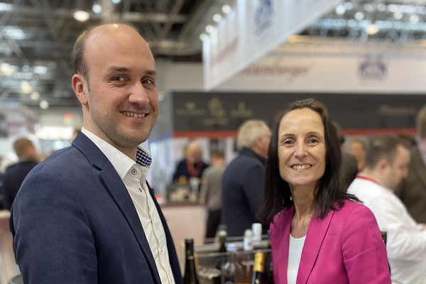 19.07._MW + Nelles Blog600 Ein Mann und eine Frau lächeln und posieren nebeneinander. Sie stehen an einem Verkaufsstand mit Getränken auf einer Messe, umgeben von anderen Personen und Ständen.