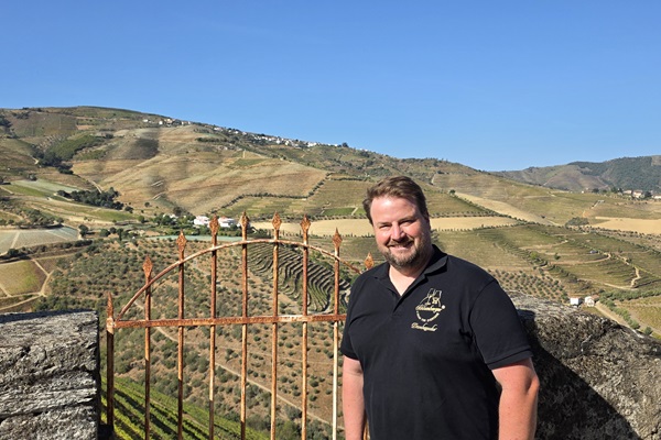 Vor einer alten, rostigen Gittertür steht ein Mann und lächelt. Im Hintergrund erstrecken sich sanfte Hügel mit Weinbergen und grünen Feldern unter einem klaren blauen Himmel.