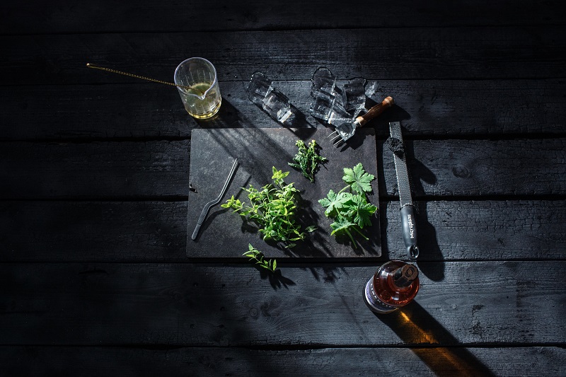 Ein schwarzes Schneidebrett liegt auf einem dunklen Tisch. Frische Kräuter, ein Bartender-Werkzeug, Eiswürfel und ein Drink sind darauf arrangiert, während Licht und Schatten interessante Kontraste schaffen.