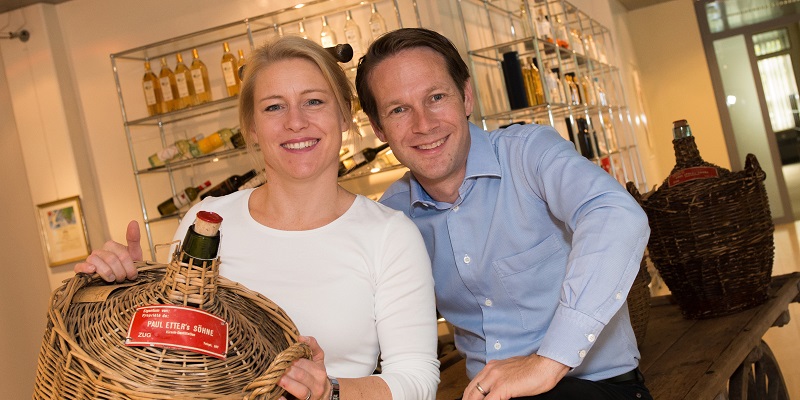 Zwei Personen lächeln in einem modernen Raum, während sie einen großen Korb mit einer Flasche in der Hand halten. Im Hintergrund stehen Regale mit Flaschen und anderen Produkten.