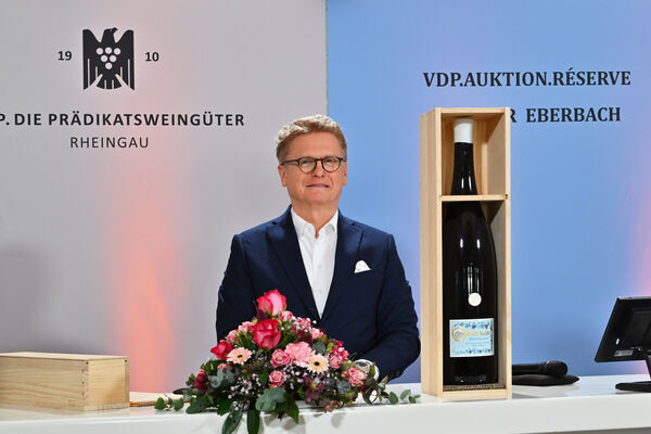 Ein Mann mit Brille steht an einem Tisch, der eine Blumenarrangement und eine Flasche in einer Holzkiste zeigt. Im Hintergrund sind Logos und Schriftzüge zu sehen, die Bezug auf Weinauktionen nehmen.