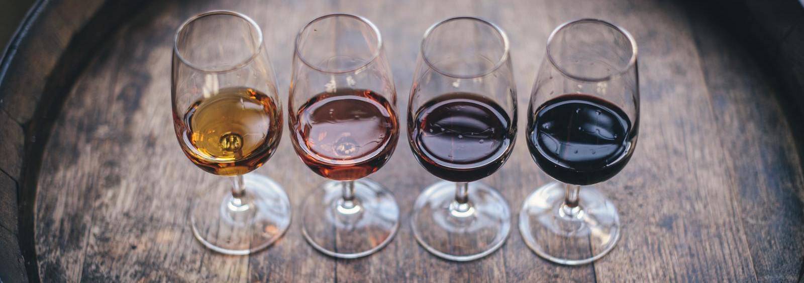 Vier Gläser mit unterschiedlichen Weinsorten stehen auf einem Holzfass. Die Weine variieren in den Farben von hellgelb bis dunkelrot, reflektieren das Licht und zeigen feine Bläschen.