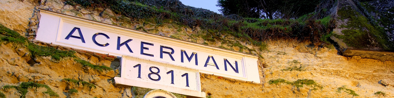 Ackerman Ein Schild mit der Aufschrift -ACKERMAN- und der Jahreszahl -1811- ist an einer naturbelassenen Wand angebracht, die vom Moos bewachsen ist, umgeben von Bäumen im Hintergrund.